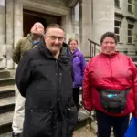 Four disability campaigners including one woman in a bright red coat and Jenny Carter wearing a purple coat. There is a man in a black coat and another man behind him with a khaki coat. They are stood on some steps and are looking at the camera.